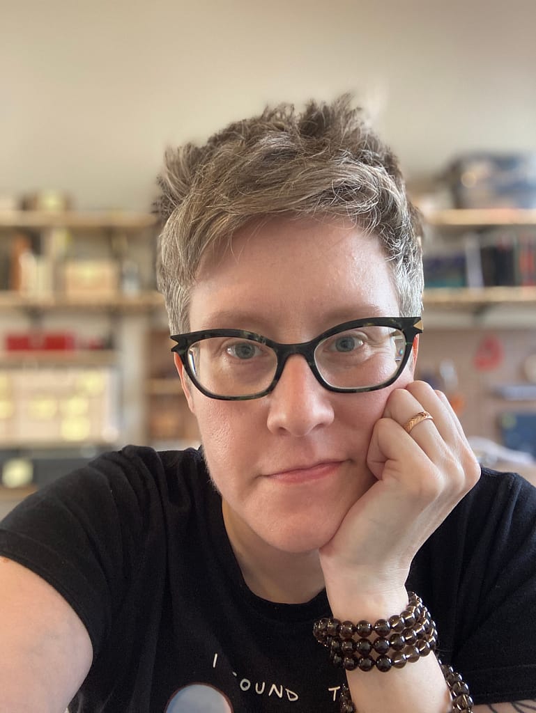 A person with short blonde hair and cat-eye glasses resting their chin in their hand. They are looking at the camera.